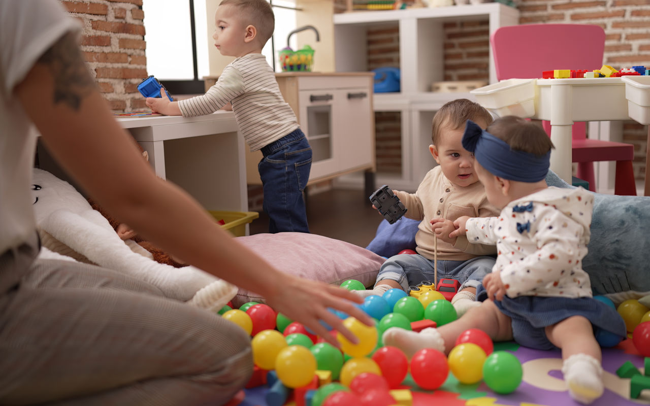 To blebørn leger på gulvet mellem små bolde - et lidt større barn står op og der sidder en voksen overfor dem