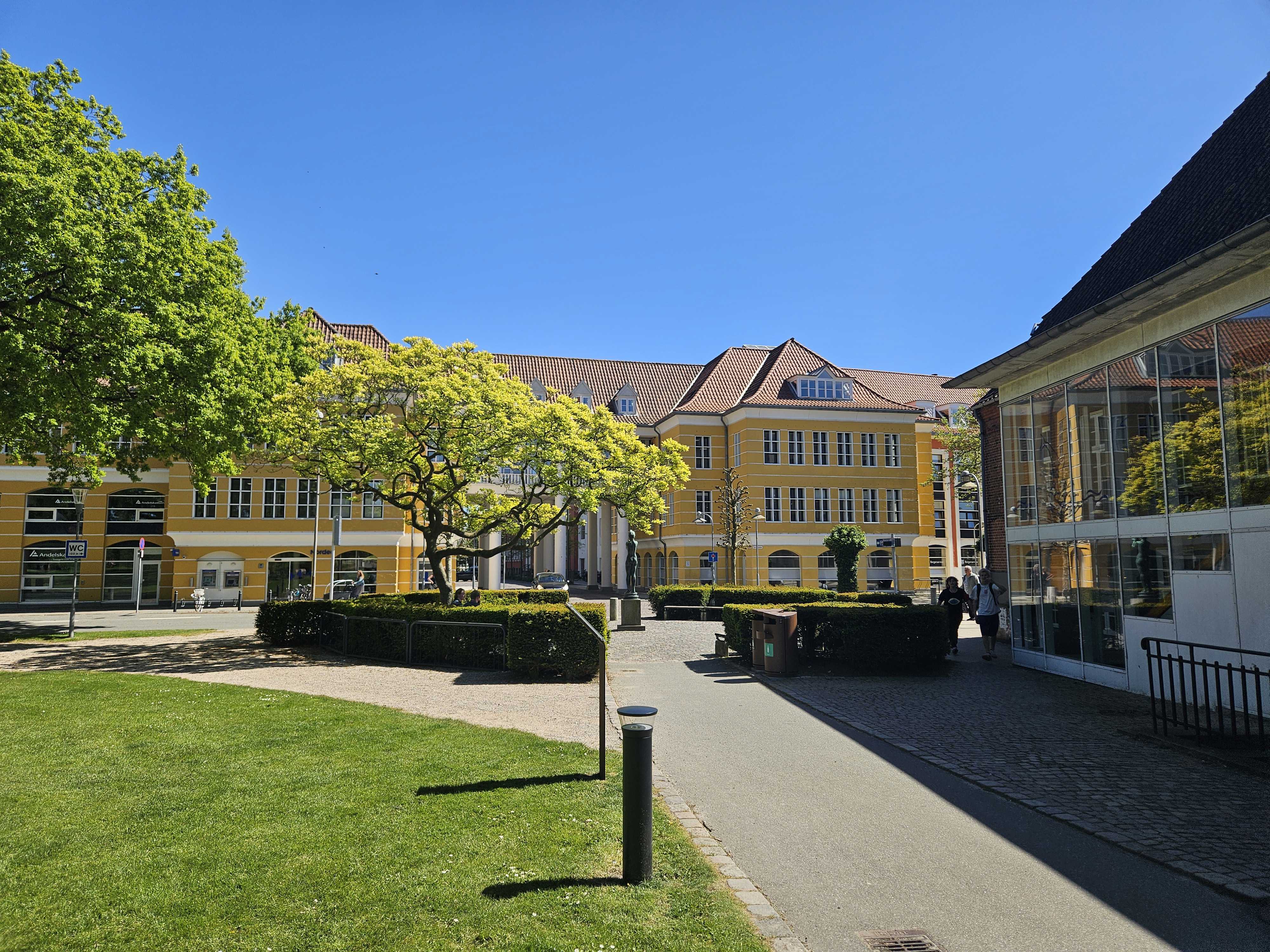 En sti med græs omkring, fører ned til en stor gul bygning med hvide vinduer. Foran bygningen er en plads omkranset af en rund hæk og et stort træ.