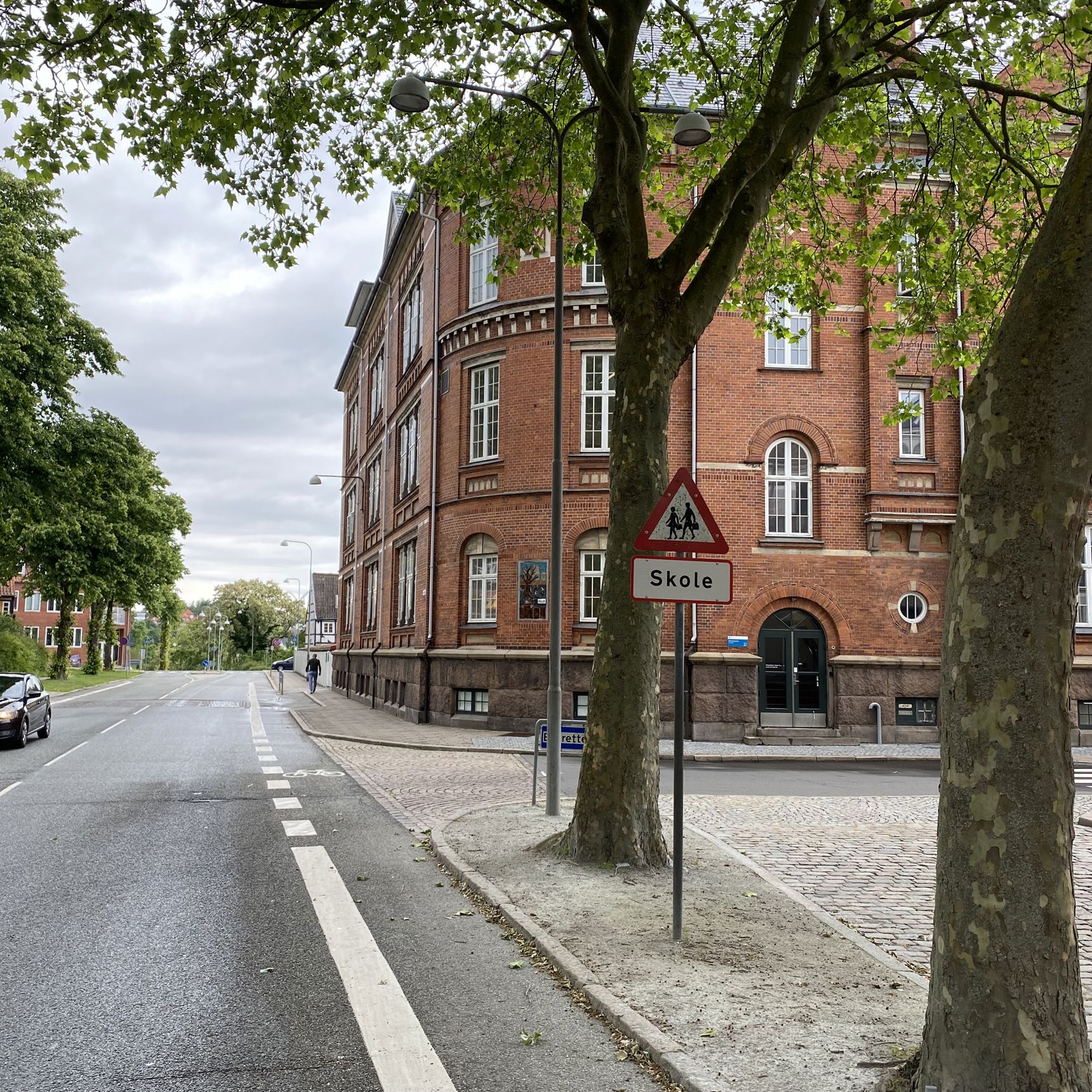 Ørkildskolen afdeling Byen - en gammel rød murstensbygning set fra vejen - udsynet er delvis blokeret med træer i forgrunden og det klassiske trekantede skilt for skole