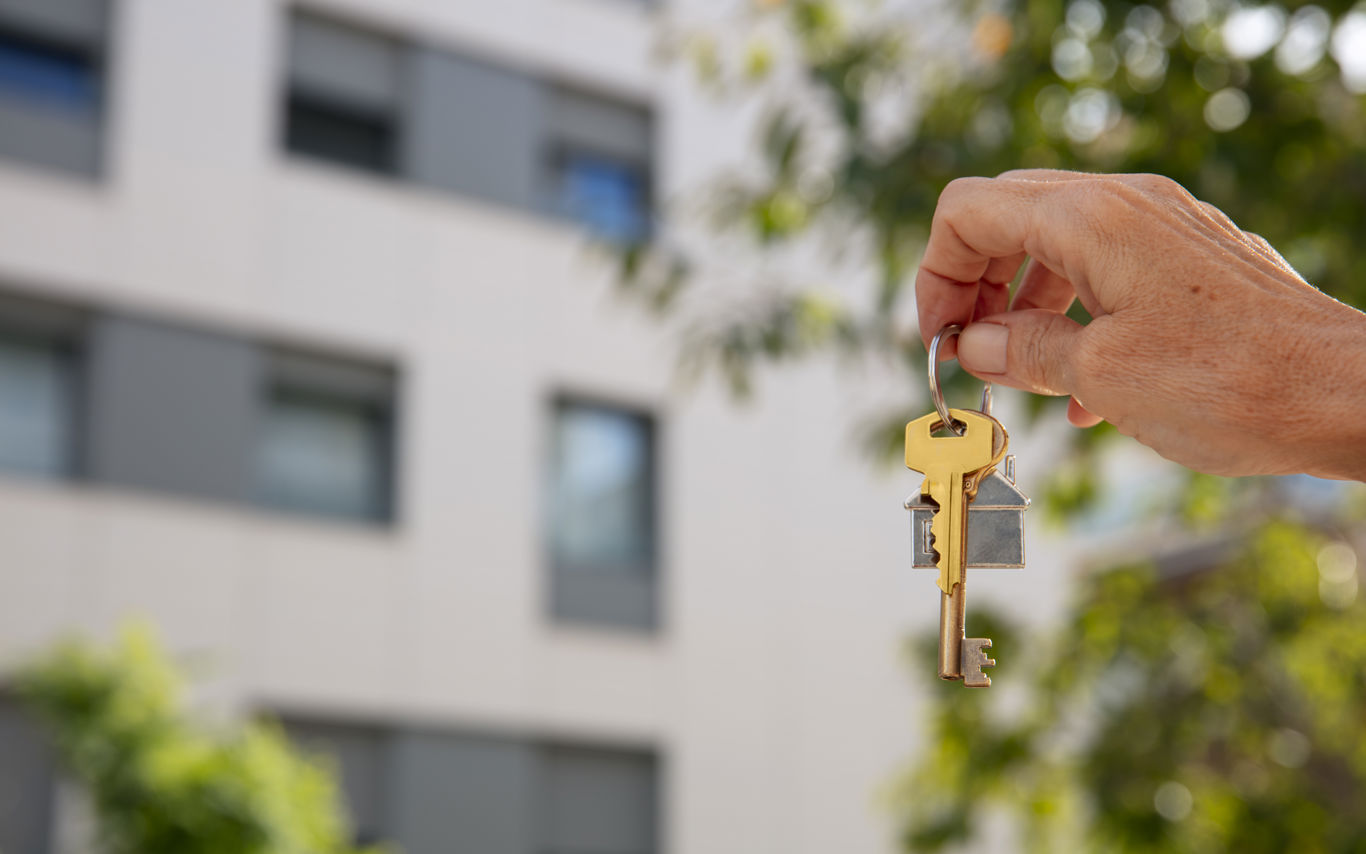 Hånd holder husnøgler. Et hus ses sløret i baggrunden