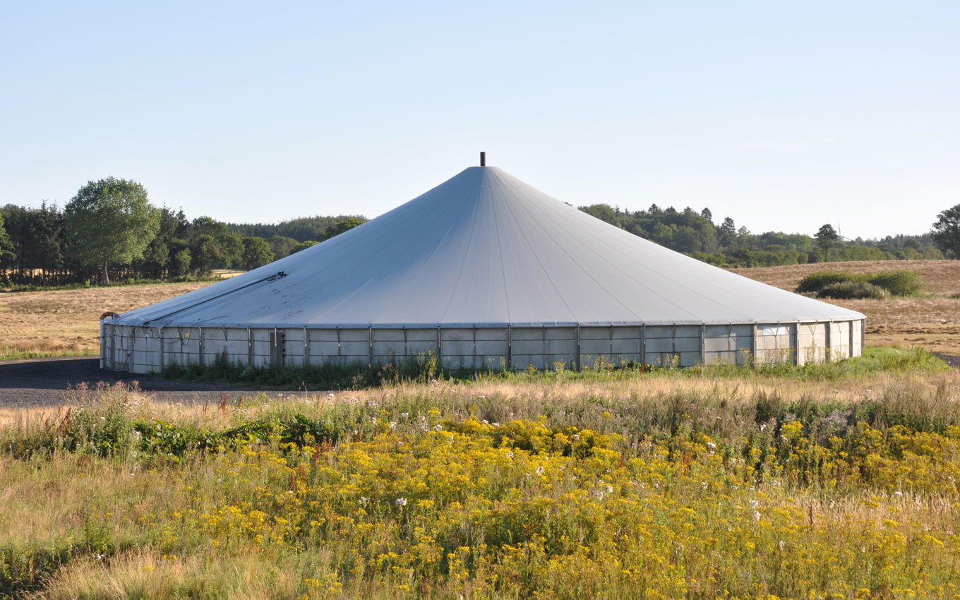 Gyllebeholder dækket med teltdug placeret i en mark med træer i baggrunden