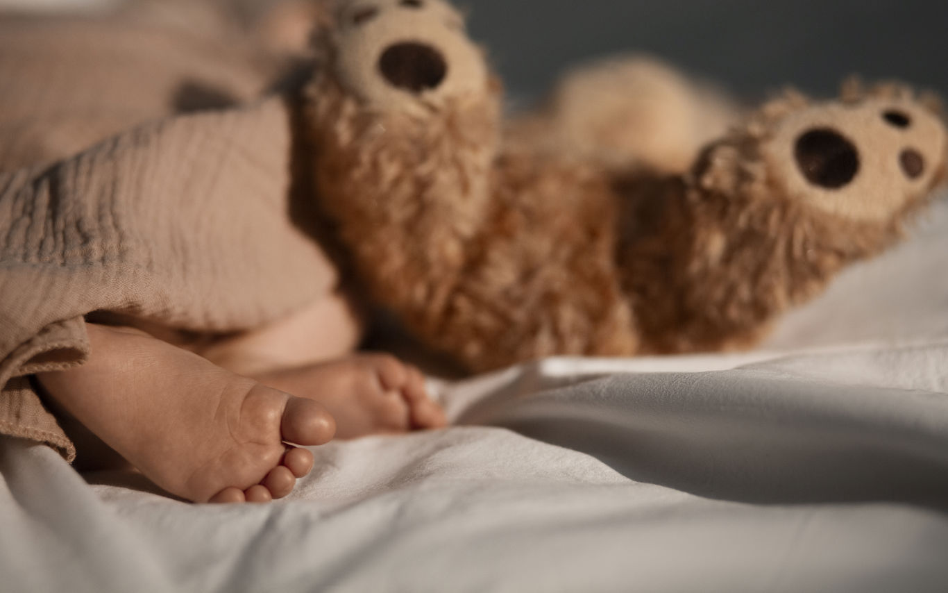 Baby fødder der stikker ud under tæppe. Brun bamses fødder ligger og stritter i vejret ved siden af baby.