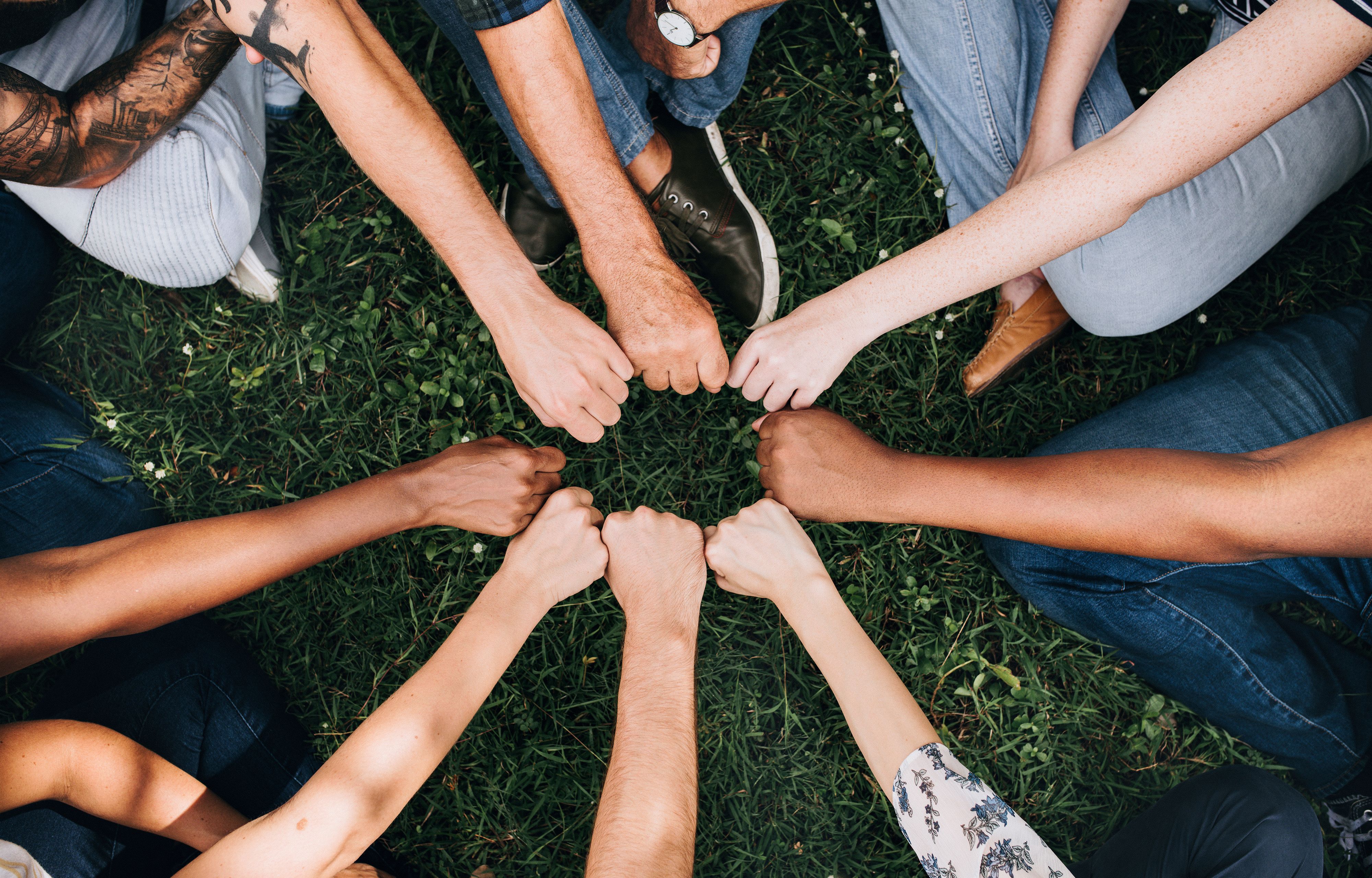 Forskellige mennesker rækker deres knyttede hænder ind mod hinanden, så de danner en cirkel. Billedet signalerer fællesskab og sammenhold.