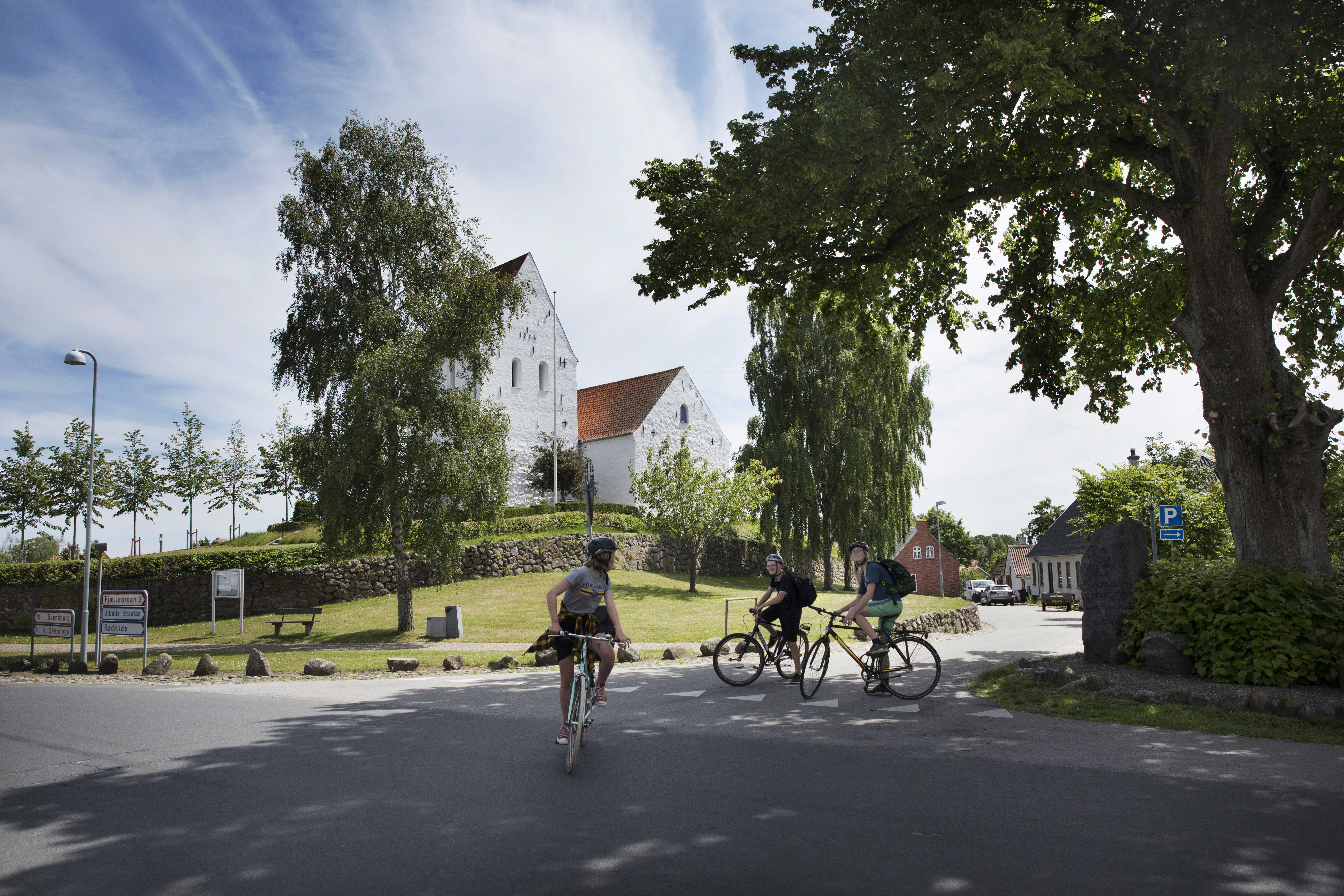 tre cykelister foran hvid kirke i grønne omgivelser
